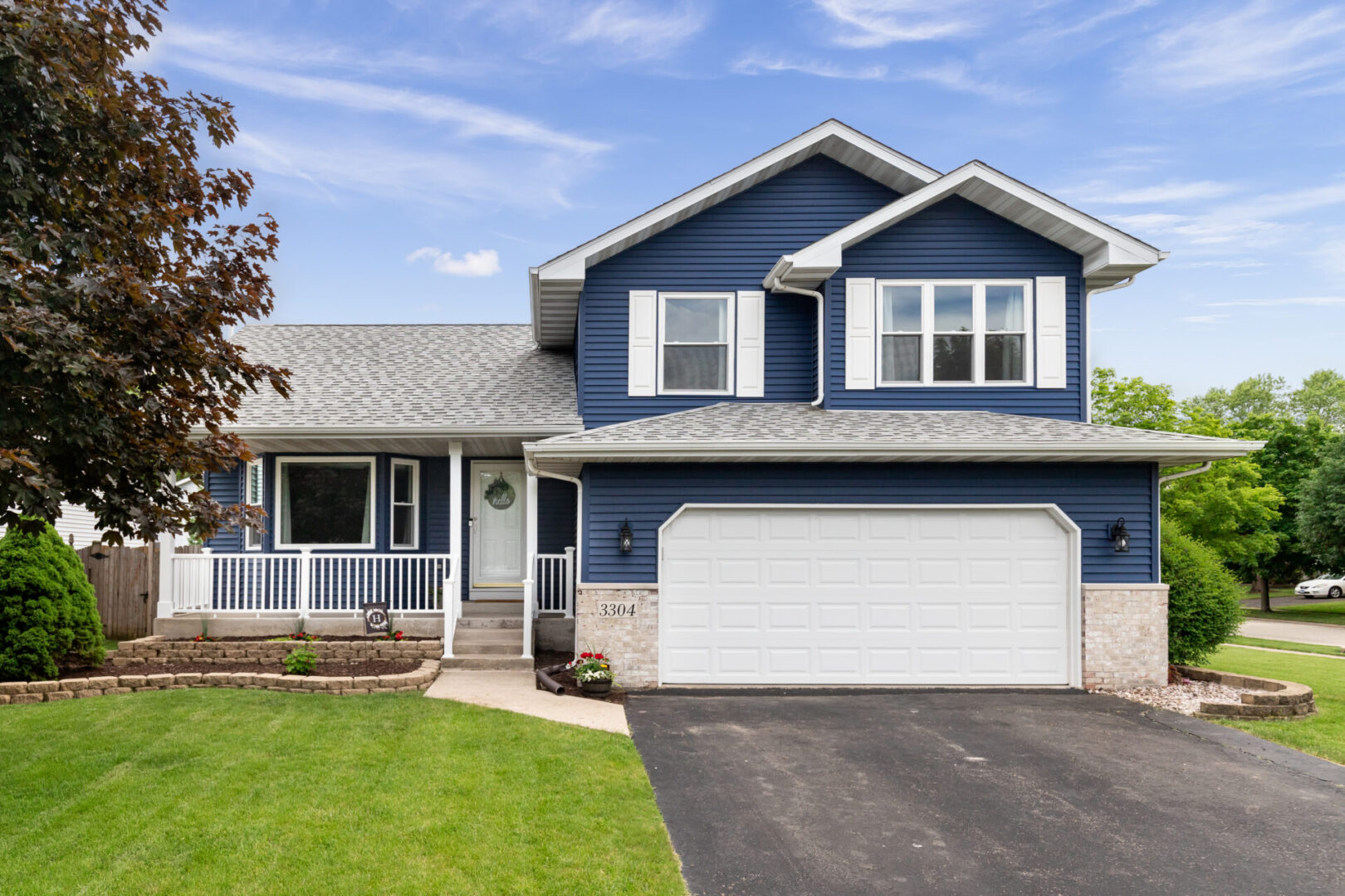 Exterior of a blue suburban home. Two-story blue house with white trim, a grey roof, and a double garage. The front yard has a lawn, a tree on the left, and a small garden bed near the entrance. Sky is partly cloudy.