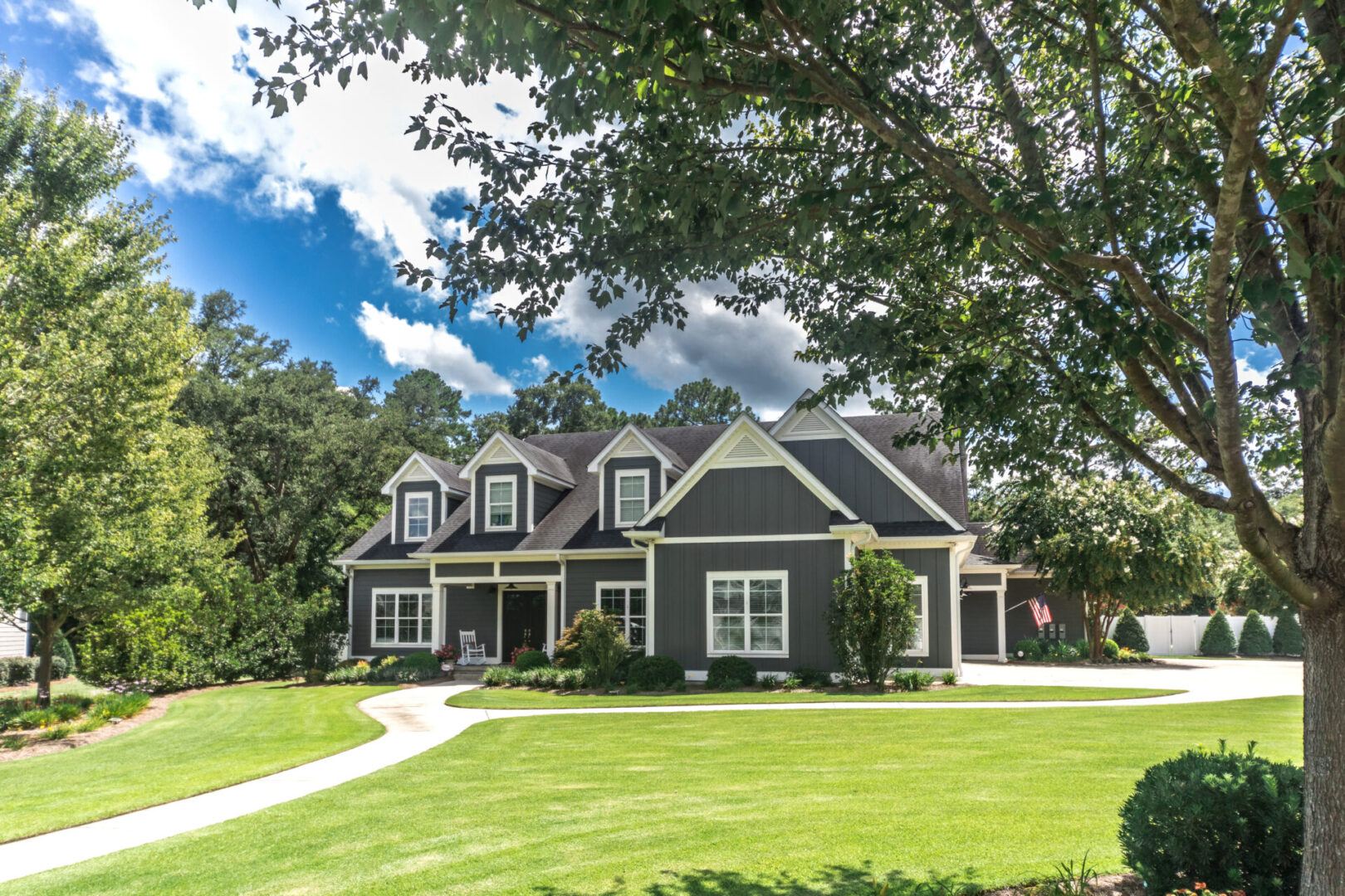A large gray craftsman new construction house with a landscaped yard and leading pathway sidewalk A two-story gray house with white trim and a gable roof, surrounded by a well-maintained lawn and trees under a partly cloudy sky.