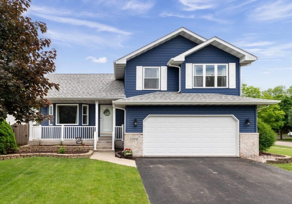 Two-story blue house with white trim, a grey roof, and a double garage. The front yard has a lawn, a tree on the left, and a small garden bed near the entrance. Sky is partly cloudy.
