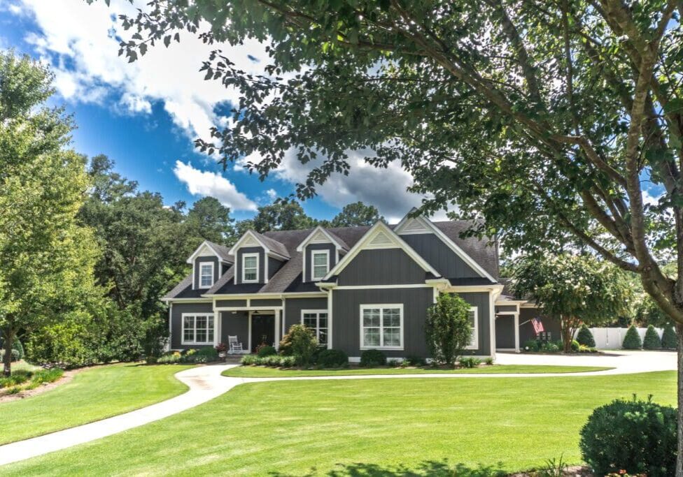 A two-story gray house with white trim and a gable roof, surrounded by a well-maintained lawn and trees under a partly cloudy sky.