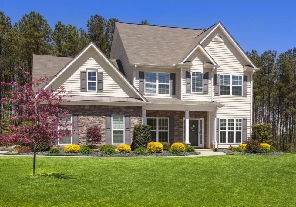 Two-story house with beige siding and stone accents, surrounded by a green lawn and shrubs, under a clear blue sky.